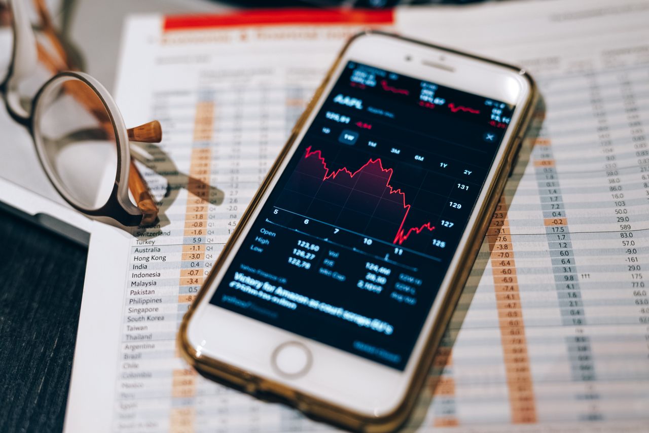 Close-up of smartphone with stock market graph on table with financial documents and glasses.